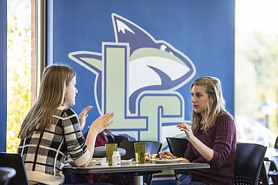 Two students at table in dining hall in front of LC Shark logo