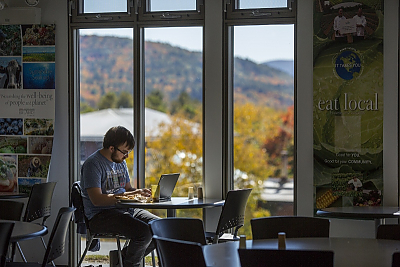 Student works at laptop in front of window in dining hall with mountains and fall foliage in background
